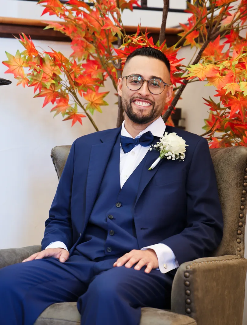 David Quintana seated in a navy three-piece suit in front of autumn foliage